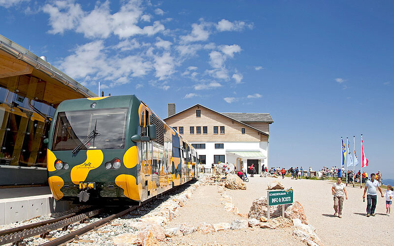 Ein grün-gelber Zug hält an einer Bergstation in der Nähe eines Holzgebäudes. Unter dem blauen Himmel gehen mehrere Menschen in der Nähe spazieren, und neben dem Gebäude sind Fahnen aufgestellt.