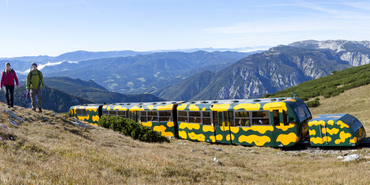 Zwei Personen wandern an einem grasbewachsenen Hang in der Nähe eines gelb-grünen Zuges entlang, während im Hintergrund Berge und Täler zu sehen sind.