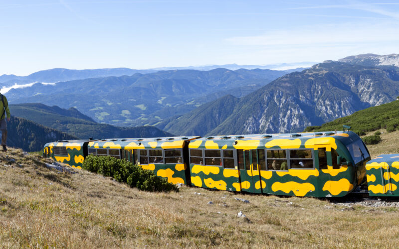 Zwei Personen wandern an einem grasbewachsenen Hang in der Nähe eines gelb-grünen Zuges entlang, während im Hintergrund Berge und Täler zu sehen sind.