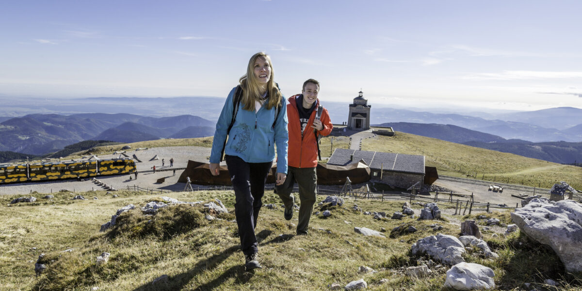 Zwei Personen wandern auf einen grasbewachsenen Hügel in der Nähe eines modernen Gebäudes und eines Aussichtsturms. Im Hintergrund sind Berge und ein geparkter gelber Zug zu sehen.