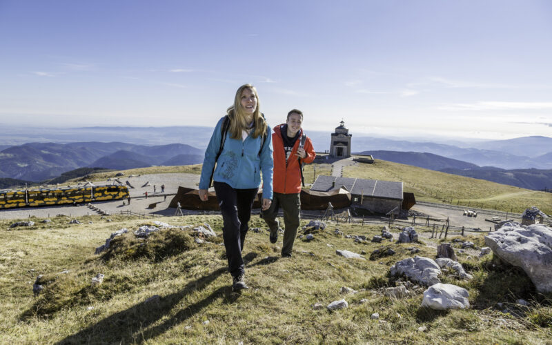 Zwei Personen wandern auf einen grasbewachsenen Hügel in der Nähe eines modernen Gebäudes und eines Aussichtsturms. Im Hintergrund sind Berge und ein geparkter gelber Zug zu sehen.