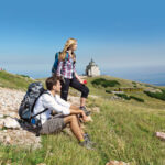 Zwei Wanderer ruhen sich mit ihren Rucksäcken auf einem grasbewachsenen Berghang aus und blicken auf ein kleines Gebäude in der Ferne unter einem klaren blauen Himmel.