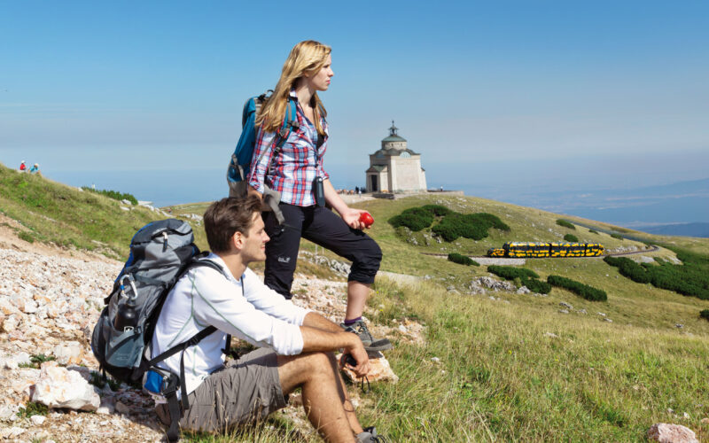 Zwei Wanderer ruhen sich mit ihren Rucksäcken auf einem grasbewachsenen Berghang aus und blicken auf ein kleines Gebäude in der Ferne unter einem klaren blauen Himmel.