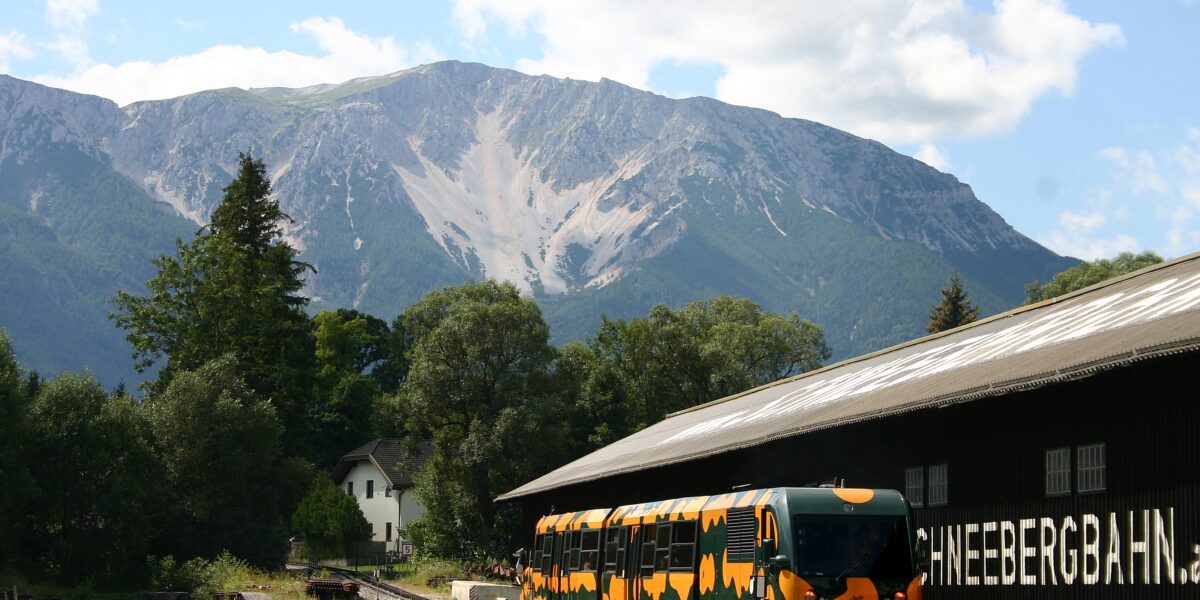Ein gelb-schwarz gemusterter Zug parkt neben einem Gebäude mit der Aufschrift "SCHNEEBERGBAHN" mit Wald und Bergen im Hintergrund.