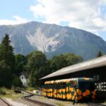 Ein gelb-schwarz gemusterter Zug parkt neben einem Gebäude mit der Aufschrift "SCHNEEBERGBAHN" mit Wald und Bergen im Hintergrund.