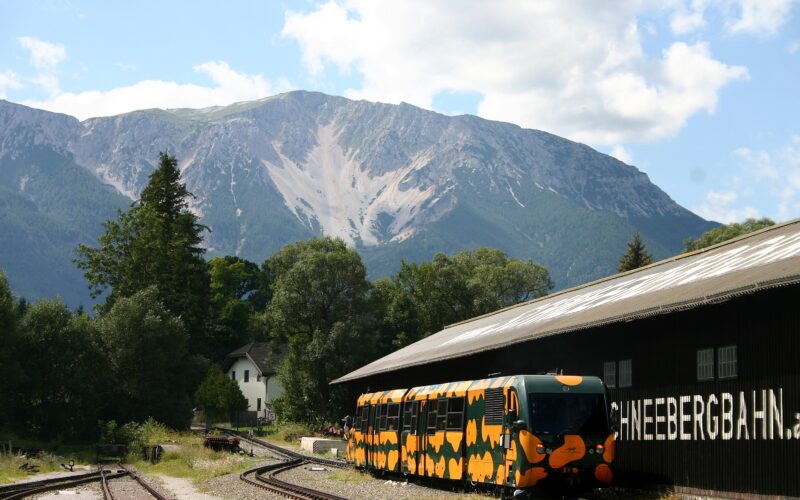 Ein gelb-schwarz gemusterter Zug parkt neben einem Gebäude mit der Aufschrift "SCHNEEBERGBAHN" mit Wald und Bergen im Hintergrund.
