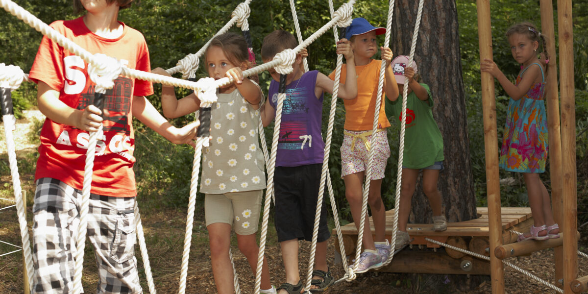 Eine Gruppe von Kindern läuft über eine Seilbrücke auf einem von Bäumen umgebenen Spielplatz.