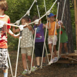 Eine Gruppe von Kindern läuft über eine Seilbrücke auf einem von Bäumen umgebenen Spielplatz.