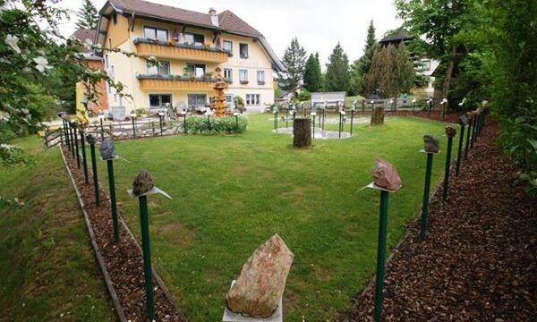 Ein grasbewachsener Garten mit Steinreihen auf Pfählen, Landschaftspflanzen, einem kleinen Springbrunnen und einem gelben Haus mit Balkonen im Hintergrund.