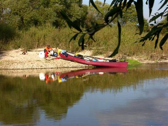 Ein rotes Kanu wird an ein Flussufer gezogen, in der Nähe befindet sich eine Campingausrüstung; eine Person sitzt neben dem Kanu am felsigen Ufer, umgeben von Bäumen und Vegetation.