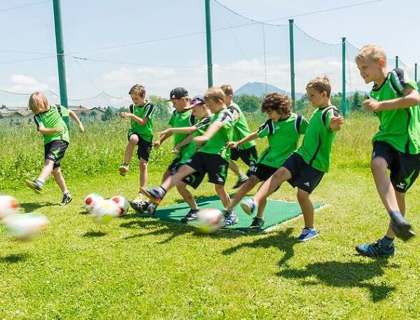 Eine Gruppe von Kindern in grünen Trikots kickt gleichzeitig Fußbälle auf einer Wiese, wobei Bewegungsunschärfe die Bälle in der Luft zeigt.