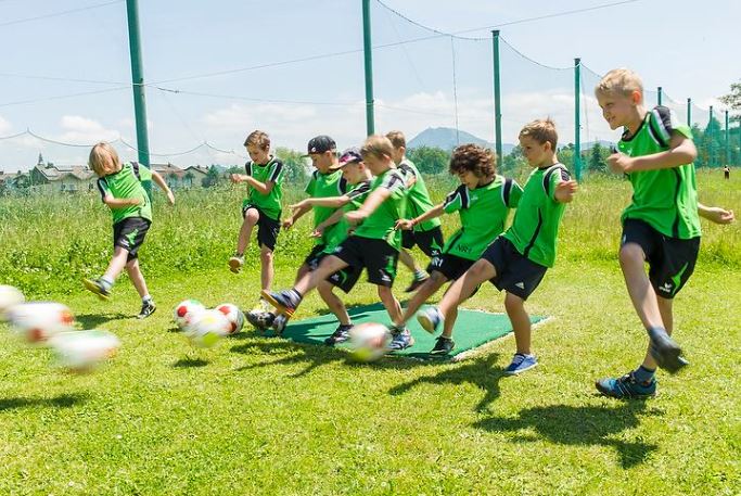 Eine Gruppe von Kindern in grünen Trikots kickt gleichzeitig Fußbälle auf einer Wiese, wobei Bewegungsunschärfe die Bälle in der Luft zeigt.