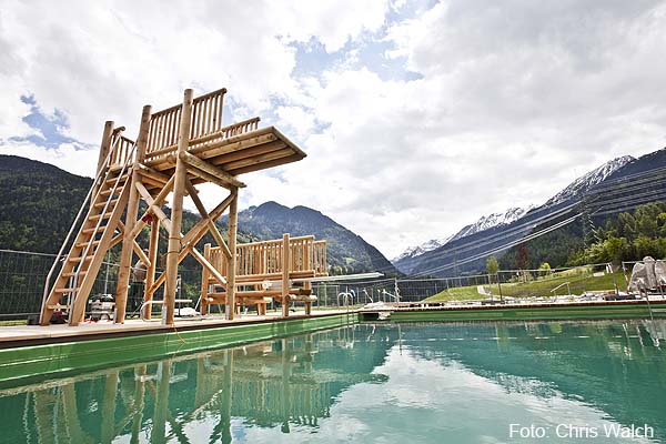 Freibad mit einem hölzernen Sprungturm, umgeben von Bergen und Grün unter einem bewölkten Himmel. Bildnachweis: Chris Walch.