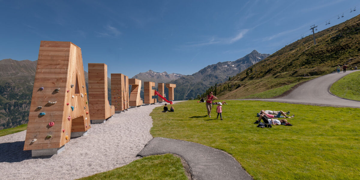 Menschen entspannen sich auf einem grasbewachsenen Hügel in der Nähe eines Spielplatzes mit Klettergerüsten in Form von großen Holzbuchstaben vor einer Berglandschaft unter einem klaren Himmel.