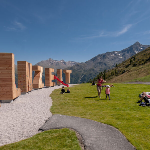 Menschen entspannen sich auf einem grasbewachsenen Hügel in der Nähe eines Spielplatzes mit Klettergerüsten in Form von großen Holzbuchstaben vor einer Berglandschaft unter einem klaren Himmel.