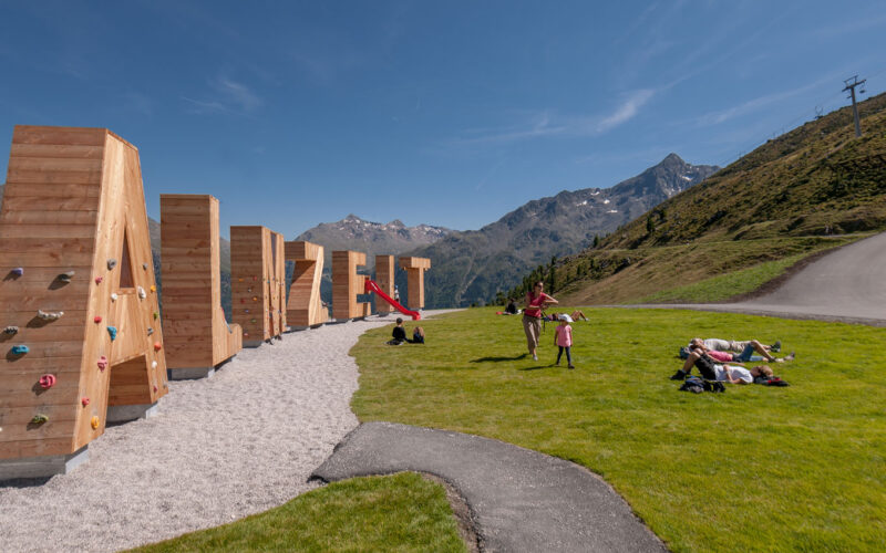 Menschen entspannen sich auf einem grasbewachsenen Hügel in der Nähe eines Spielplatzes mit Klettergerüsten in Form von großen Holzbuchstaben vor einer Berglandschaft unter einem klaren Himmel.