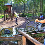 Kinder spielen auf einem Waldspielplatz mit Rutschen und einem Wasserspiel; im Vordergrund bedient ein Mädchen in einem roten Hemd eine Wasserpumpe.