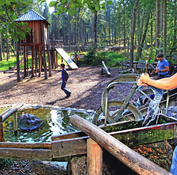Kinder spielen auf einem Waldspielplatz mit Rutschen und einem Wasserspiel; im Vordergrund bedient ein Mädchen in einem roten Hemd eine Wasserpumpe.