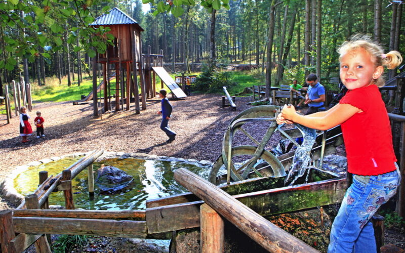 Kinder spielen auf einem Waldspielplatz mit Rutschen und einem Wasserspiel; im Vordergrund bedient ein Mädchen in einem roten Hemd eine Wasserpumpe.