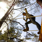 Eine Person, die einen Helm und einen Klettergurt trägt, läuft auf Seilen zwischen Bäumen in einem Outdoor-Abenteuerpark, wobei das Sonnenlicht durch die Äste sichtbar ist.
