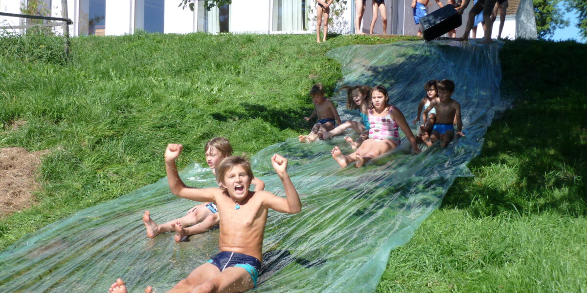 Kinder rutschen eine selbstgebaute Wasserrutsche auf einem Grashügel hinunter, oben wartet eine Gruppe und im Hintergrund ist ein weißes Gebäude zu sehen.