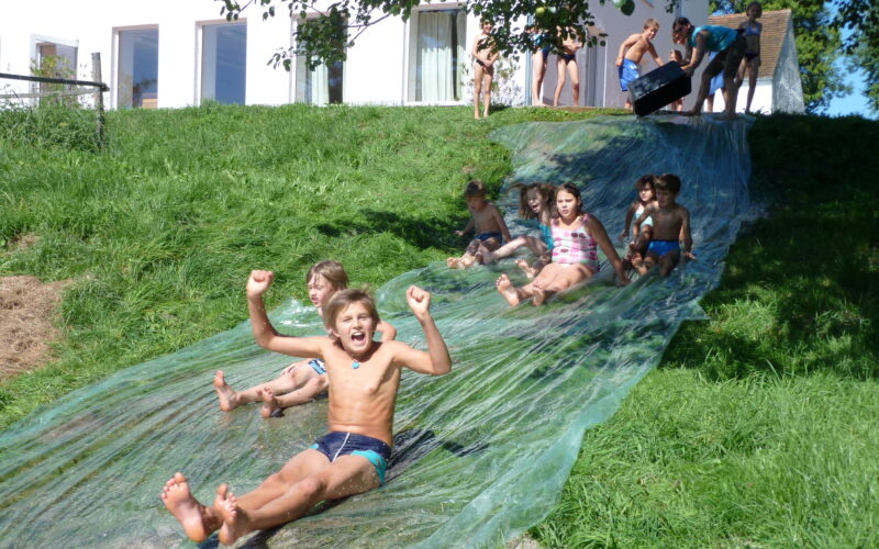 Kinder rutschen eine selbstgebaute Wasserrutsche auf einem Grashügel hinunter, oben wartet eine Gruppe und im Hintergrund ist ein weißes Gebäude zu sehen.