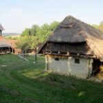 Ein reetgedecktes Haus und ein hölzerner Glockenturm stehen auf einer Wiese, die von Bäumen und einem Holzzaun umgeben ist.
