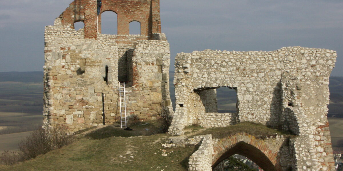 Die steinernen Ruinen einer alten Burg oder Festung liegen auf einem grasbewachsenen Hügel unter einem wolkenverhangenen Himmel, eine Metallleiter lehnt an der Wand, und im Hintergrund sind weite Felder zu sehen.