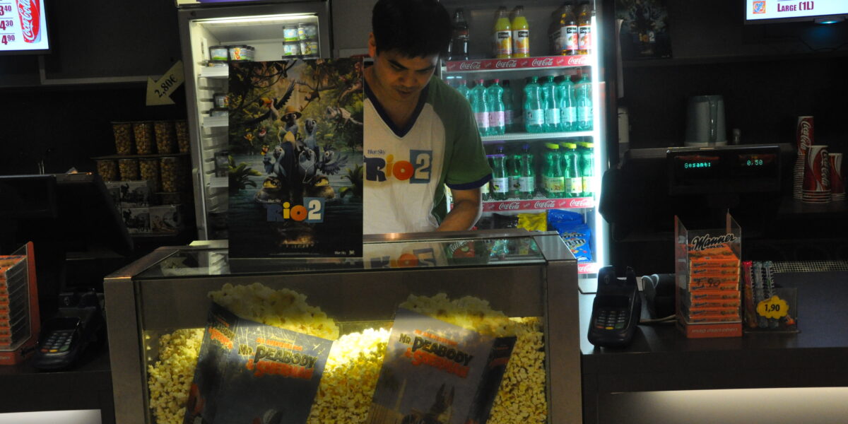 Ein Angestellter bereitet Popcorn hinter einer Glasvitrine mit Filmplakaten zu, während im Hintergrund Getränke und Snacks zu sehen sind.