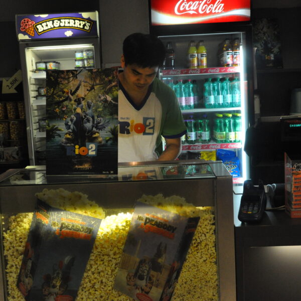 Ein Angestellter bereitet Popcorn hinter einer Glasvitrine mit Filmplakaten zu, während im Hintergrund Getränke und Snacks zu sehen sind.