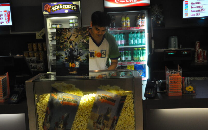 Ein Angestellter bereitet Popcorn hinter einer Glasvitrine mit Filmplakaten zu, während im Hintergrund Getränke und Snacks zu sehen sind.