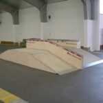 Eine hölzerne Skateboard-Rampenstruktur mit einer schrägen Oberfläche und Metallkanten befindet sich in einer Halle mit Betonboden.
