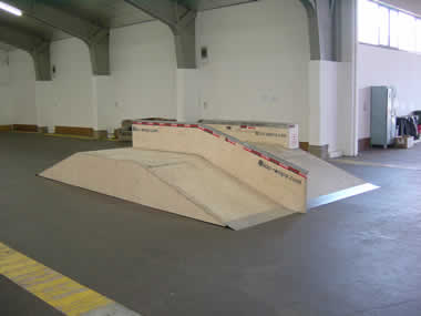 Eine hölzerne Skateboard-Rampenstruktur mit einer schrägen Oberfläche und Metallkanten befindet sich in einer Halle mit Betonboden.