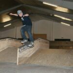 Ein Skateboarder, der eine Mütze trägt, führt einen Trick auf einer Kante in einem Indoor-Skatepark aus.