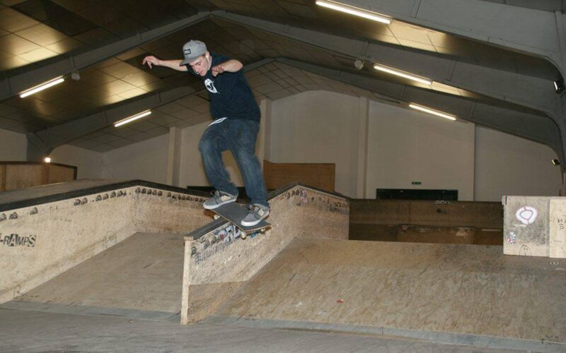 Ein Skateboarder, der eine Mütze trägt, führt einen Trick auf einer Kante in einem Indoor-Skatepark aus.