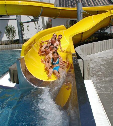 Eine Gruppe von Kindern in Badeanzügen rutscht eine gelbe Wasserrutsche hinunter in ein Becken in einem Freibad.