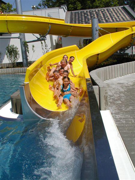 Eine Gruppe von Kindern in Badeanzügen rutscht eine gelbe Wasserrutsche hinunter in ein Becken in einem Freibad.