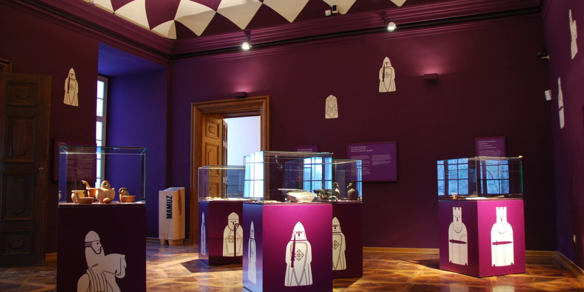 Ein Museumsraum mit violetten Wänden und einer violetten Decke, Glasvitrinen mit Artefakten und weißen schachähnlichen Illustrationen auf den Sockeln und an den Wänden.