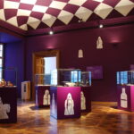 Ein Museumsraum mit violetten Wänden und einer violetten Decke, Glasvitrinen mit Artefakten und weißen schachähnlichen Illustrationen auf den Sockeln und an den Wänden.