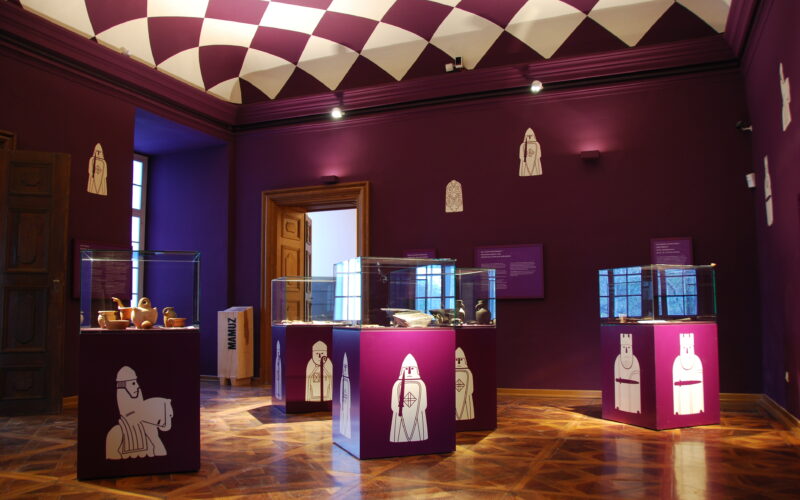 Ein Museumsraum mit violetten Wänden und einer violetten Decke, Glasvitrinen mit Artefakten und weißen schachähnlichen Illustrationen auf den Sockeln und an den Wänden.