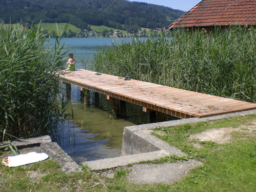 Ein hölzerner Steg erstreckt sich über klares Wasser, umgeben von Schilf, an dessen Ende eine Person in der Nähe eines Sees und eines rotgedeckten Gebäudes sitzt.