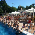 Menschenmenge an einem Freibad, einige sitzen am Rand und andere stehen oder gehen in der Nähe; weiße Zelte und Bäume im Hintergrund an einem sonnigen Tag.