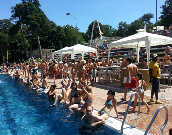 Menschenmenge an einem Freibad, einige sitzen am Rand und andere stehen oder gehen in der Nähe; weiße Zelte und Bäume im Hintergrund an einem sonnigen Tag.