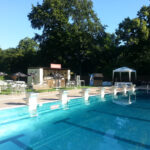 Freibad mit Sprungtürmen im Vordergrund, umgeben von Bäumen, Tischen, Sonnenschirmen und einem kleinen Erfrischungsstand im Hintergrund unter strahlend blauem Himmel.