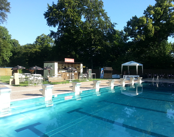 Freibad mit Sprungtürmen im Vordergrund, umgeben von Bäumen, Tischen, Sonnenschirmen und einem kleinen Erfrischungsstand im Hintergrund unter strahlend blauem Himmel.