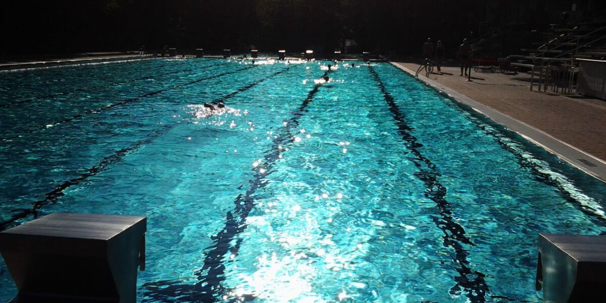Freibad mit mehreren Bahnen, Sonnenlicht, das sich auf dem Wasser spiegelt, Startblöcke im Vordergrund und ein paar Leute auf dem Pooldeck im Hintergrund.