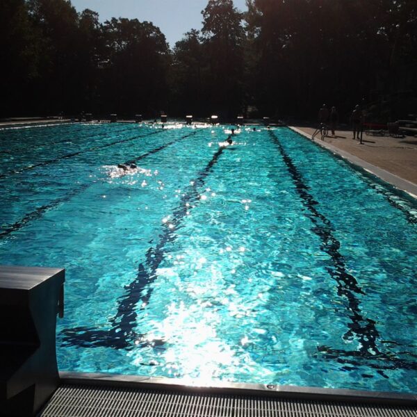 Freibad mit mehreren Bahnen, Sonnenlicht, das sich auf dem Wasser spiegelt, Startblöcke im Vordergrund und ein paar Leute auf dem Pooldeck im Hintergrund.