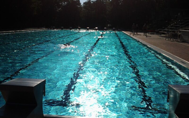 Freibad mit mehreren Bahnen, Sonnenlicht, das sich auf dem Wasser spiegelt, Startblöcke im Vordergrund und ein paar Leute auf dem Pooldeck im Hintergrund.