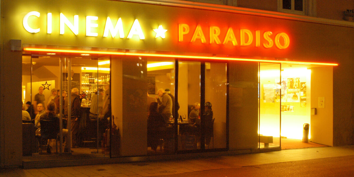 Außenansicht des Cinema Paradiso bei Nacht, mit Menschen, die durch die großen Fenster und die gelbe und rote Leuchtreklame über dem Eingang ins Innere gelangen.