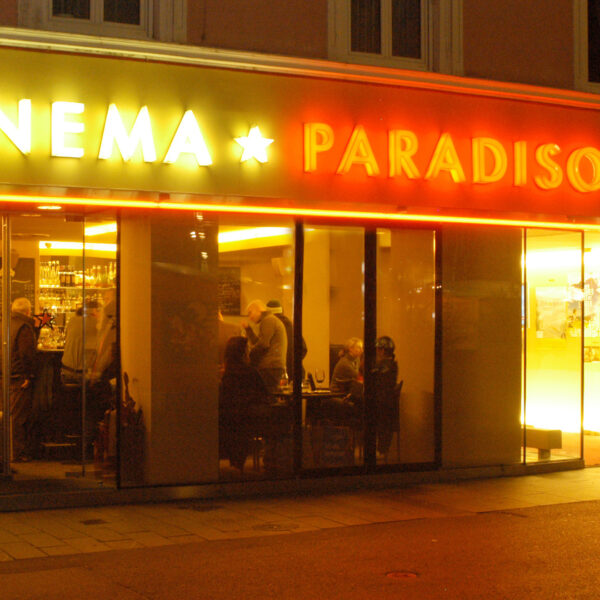 Außenansicht des Cinema Paradiso bei Nacht, mit Menschen, die durch die großen Fenster und die gelbe und rote Leuchtreklame über dem Eingang ins Innere gelangen.